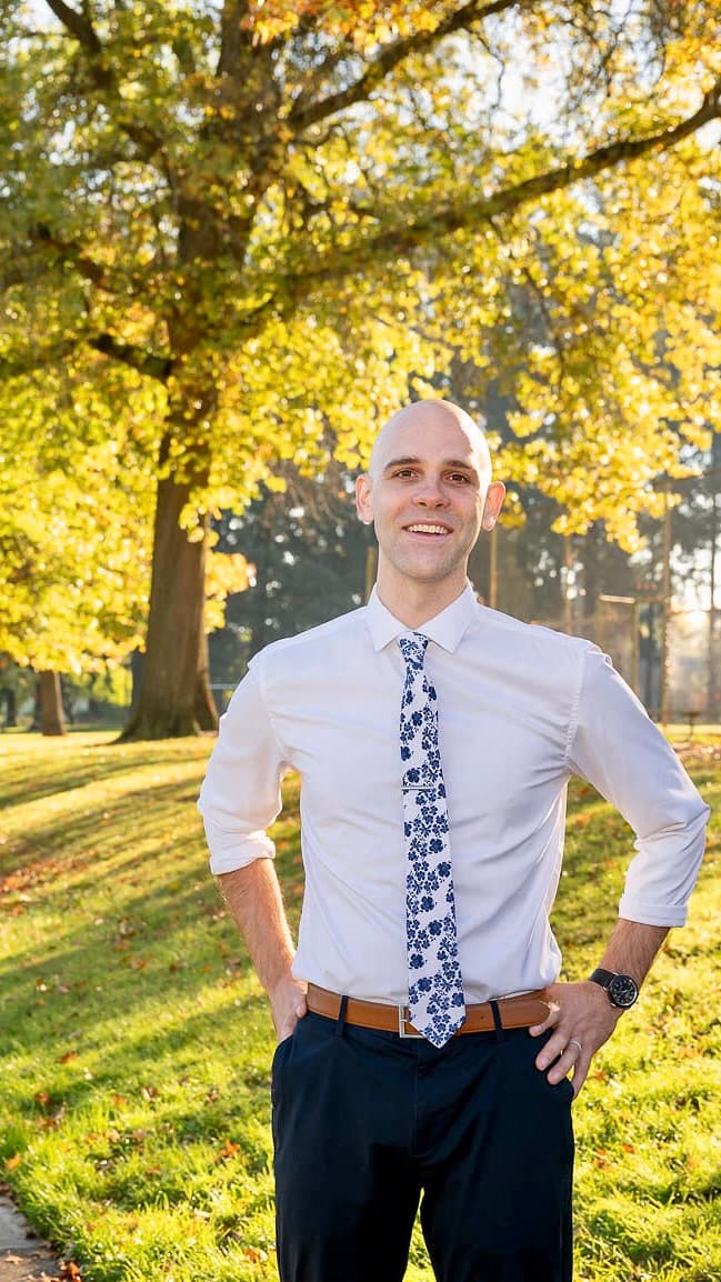 Joseph Emerson in a white shirt with a floral blue tie and blue trousers stands with his hands on his hips. He is wearing a brown belt and a watch on his left wrist. The background is a sunlit park with tall trees bearing golden autumn leaves, and a well-kept lawn scattered with fallen leaves, suggesting a bright autumn day