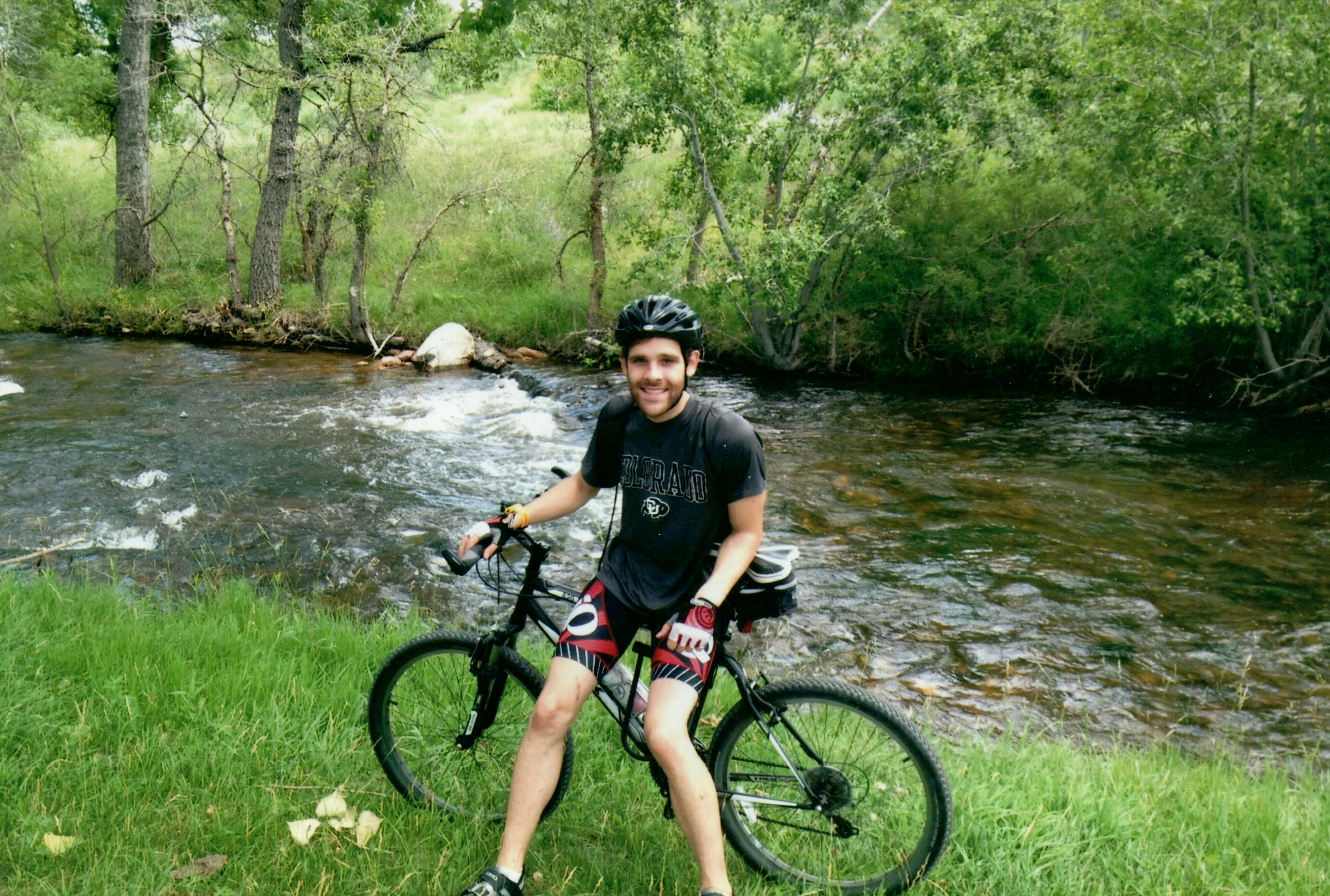 Joseph Emerson with a beard, wearing a black T-shirt, cycling shorts, and a bicycle helmet, stands straddling a mountain bike beside a flowing river. The river is surrounded by lush greenery and trees, with some rocks visible in the water.