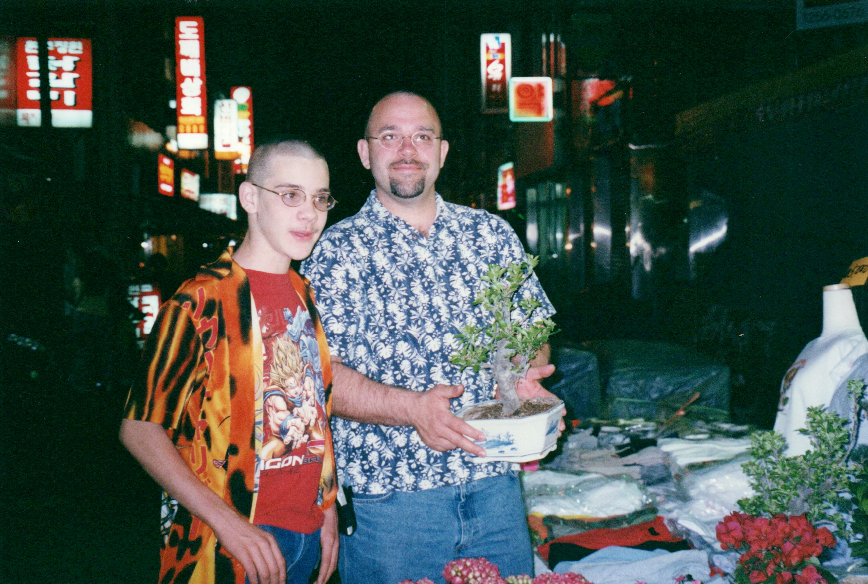 Joseph Emerson with his father in front of a Korean night market bustling with neon signboards. On the left, Joseph sports a laughable flame-patterned shirt over a Dragon Ball Z tee. To the right, Dad holds a small bonsai tree. The foreground shows a variety of goods on display, suggesting a vibrant street market scene.